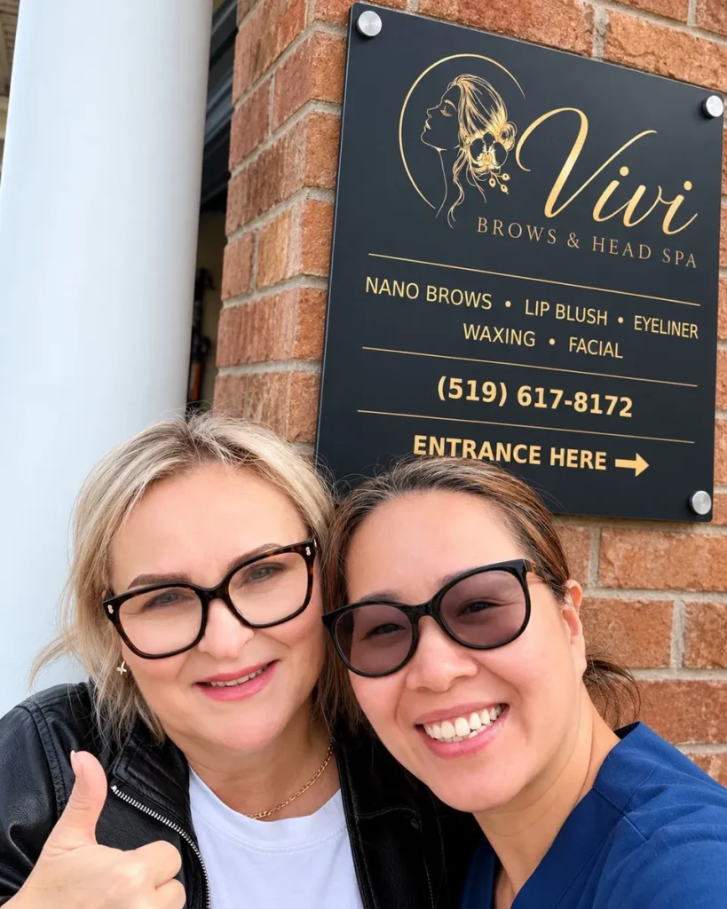 Smiling client giving a thumbs-up beside the artist under the Vivi Brows & Head Spa sign in London, Ontario